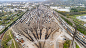 A drone shot captures the vast expanse of the railroad yard in Chattanooga, showcasing the intricate network of tracks stretching out in all directions. The lines of trains, freight cars, and locomotives create a captivating scene of organized chaos, with the city's industrial heart visible in the background. The overhead perspective highlights the scale of the operation, with trains moving along the tracks and creating a sense of constant motion. The juxtaposition of steel and the surrounding natural landscape of the Tennessee River provides a stunning contrast in this industrial yet scenic view of Chattanooga.