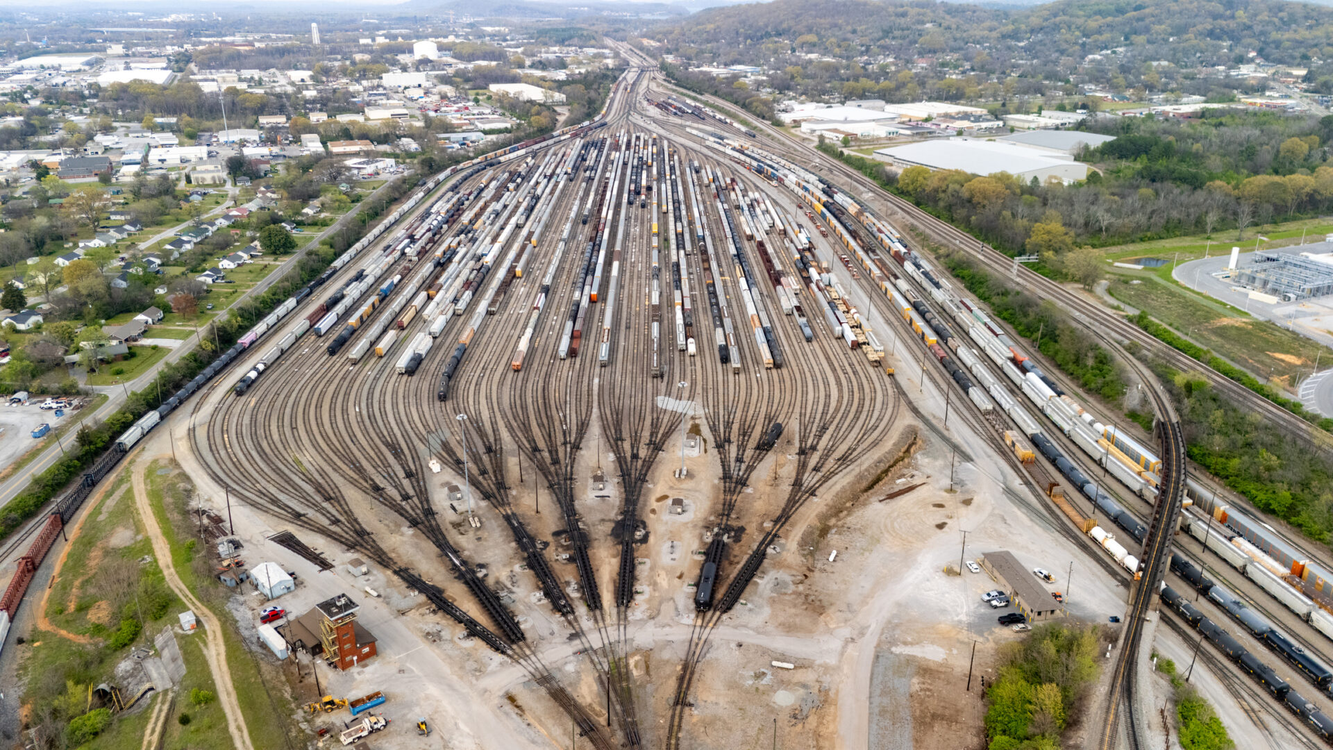 A drone shot captures the vast expanse of the railroad yard in Chattanooga, showcasing the intricate network of tracks stretching out in all directions. The lines of trains, freight cars, and locomotives create a captivating scene of organized chaos, with the city's industrial heart visible in the background. The overhead perspective highlights the scale of the operation, with trains moving along the tracks and creating a sense of constant motion. The juxtaposition of steel and the surrounding natural landscape of the Tennessee River provides a stunning contrast in this industrial yet scenic view of Chattanooga.